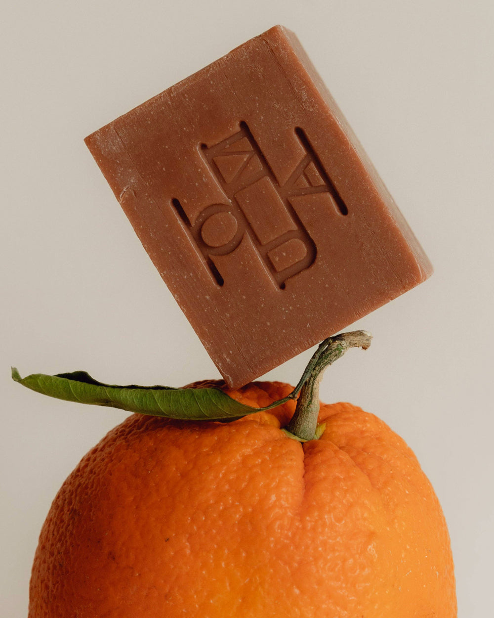 A rectangular bar of brown soap with an embossed monogram that reads OUMA, balanced on top of an orange with a green leaf, against a light gray background.
