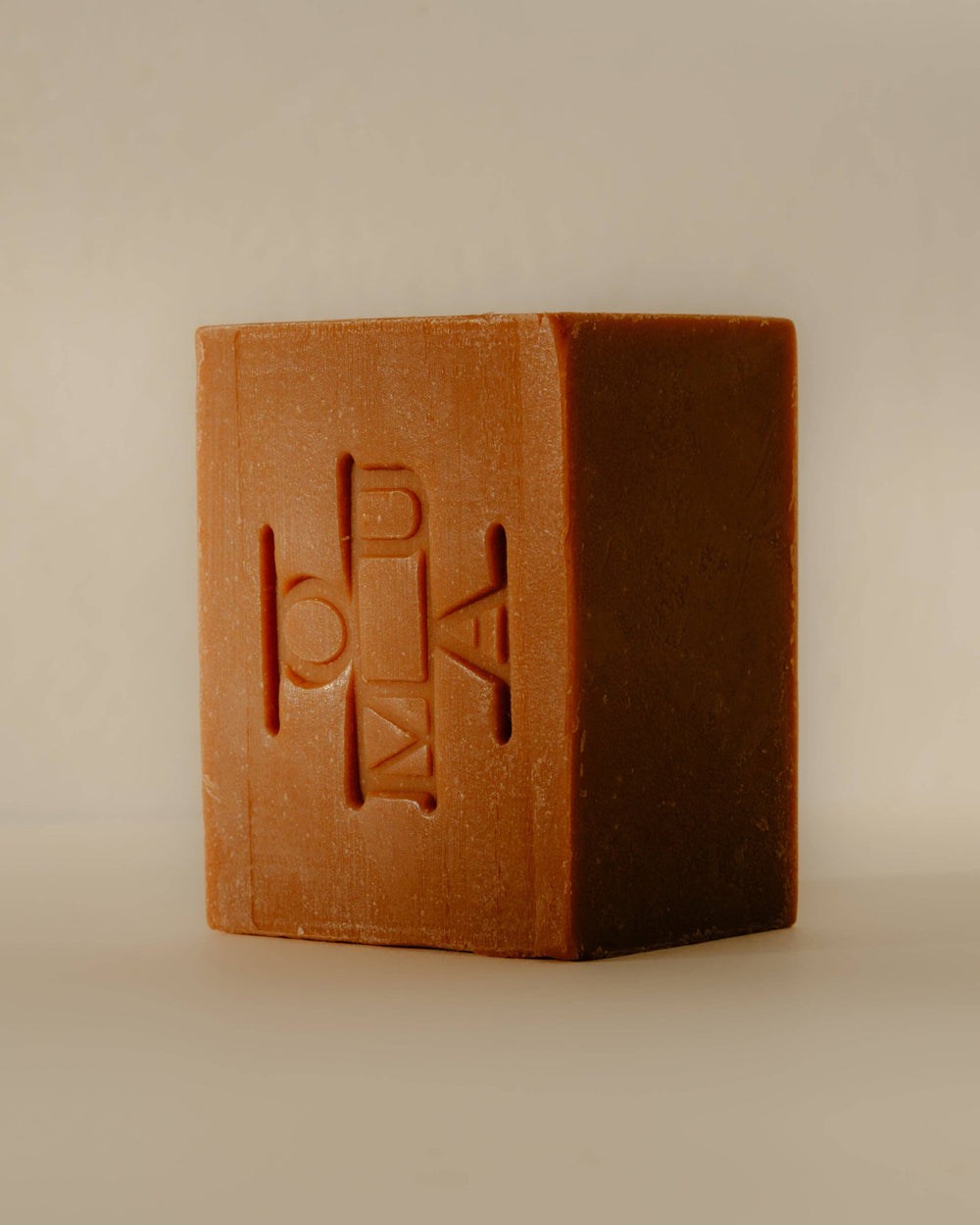 A rectangular bar of brown soap with an embossed logo that reads OUMA, standing upright against a plain beige background.