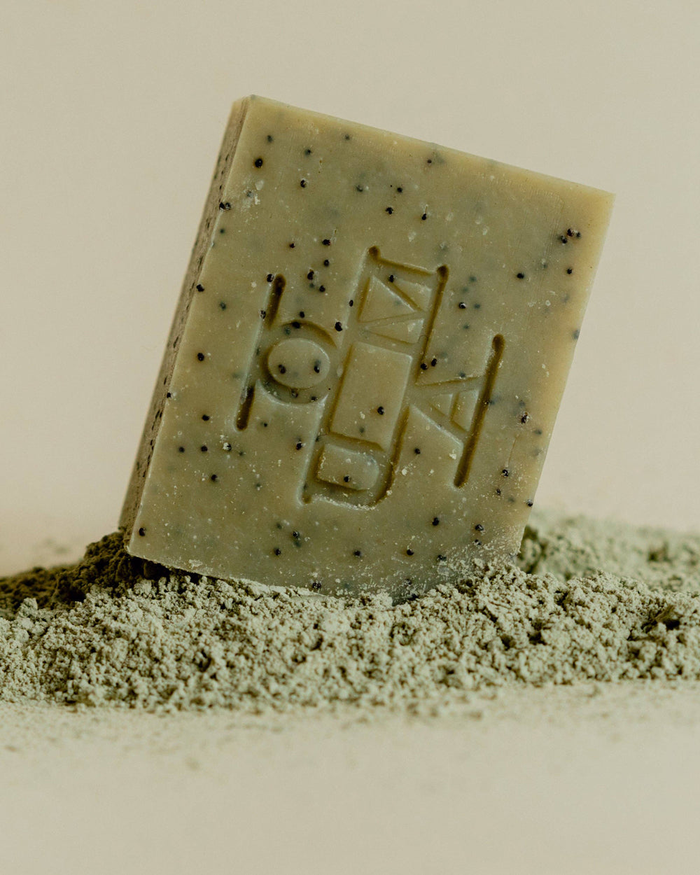 A rectangular bar of soap standing upright on a small pile of fine powder, photographed against a plain light beige background. The soap has a muted sage-green tone with scattered dark specks and an embossed geometric logo on the front that reads OUMA.