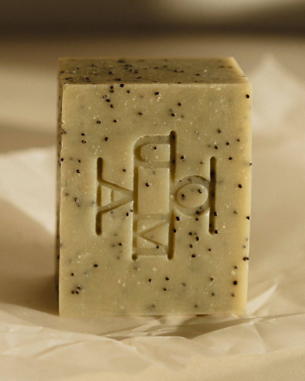 A rectangular bar of soap standing upright  and photographed against a light beige background. The soap has a muted sage-green tone with scattered dark specks and an embossed geometric logo on the front that reads OUMA.