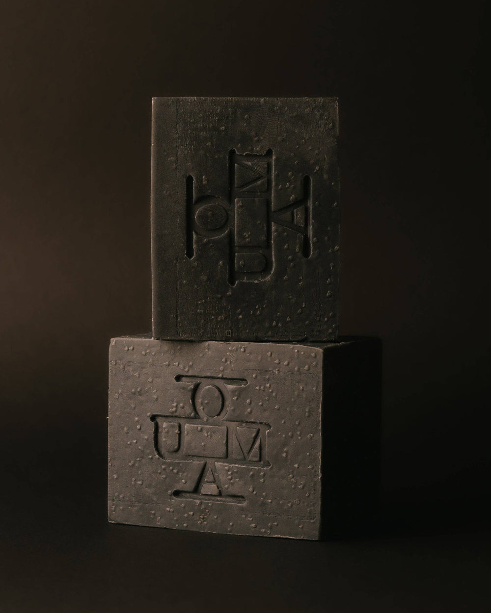 Two dark black textured rectangular blocks of soap with embossed geometric logos that read OUMA, photographed on a warm beige background.