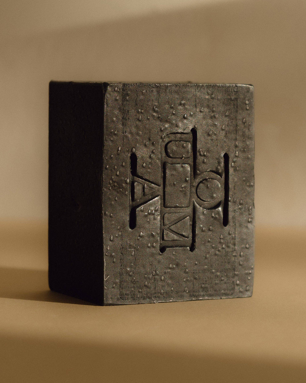 A dark black textured rectangular block of soap with an embossed geometric logo that reads OUMA, photographed on a warm beige background.