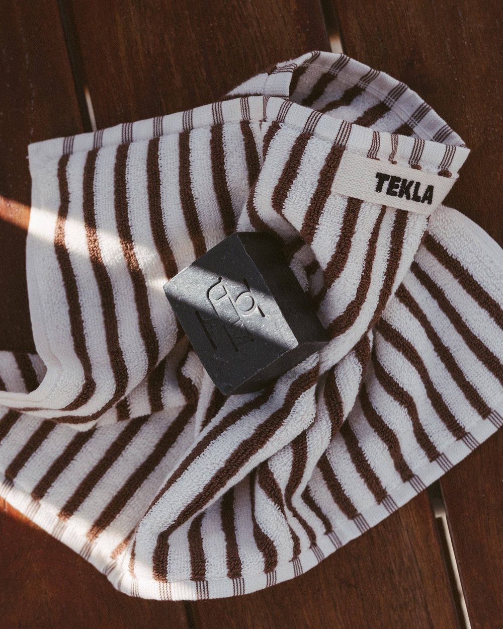 A striped terrycloth towel with dark brown and white vertical stripes, featuring a white woven label with the brand name TEKLA. A dark gray soap bar is draped over the towel, which is laid out on a dark wooden surface.