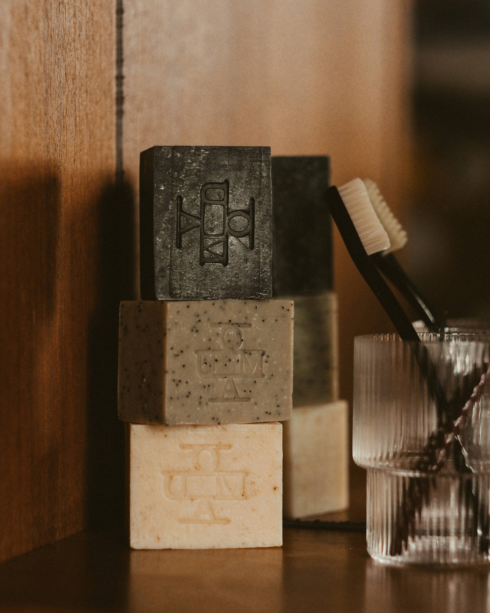 Three stacked rectangular bars of soap in dark charcoal, speckled taupe, and light beige, each embossed with a monogram logo that reads OUMA, displayed on a wooden surface next to a glass cup holding toothbrushes.