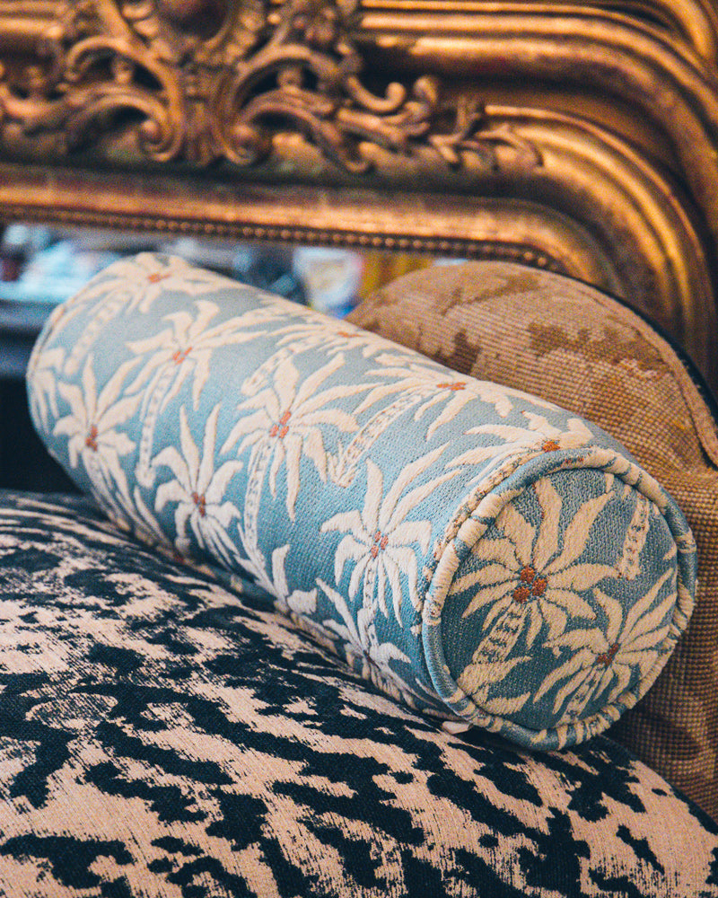 An elegant bolster pillow by Dutch brand Palais, featuring soft blue tones and white palm tree patterns, set against a backdrop of antique mirror details.