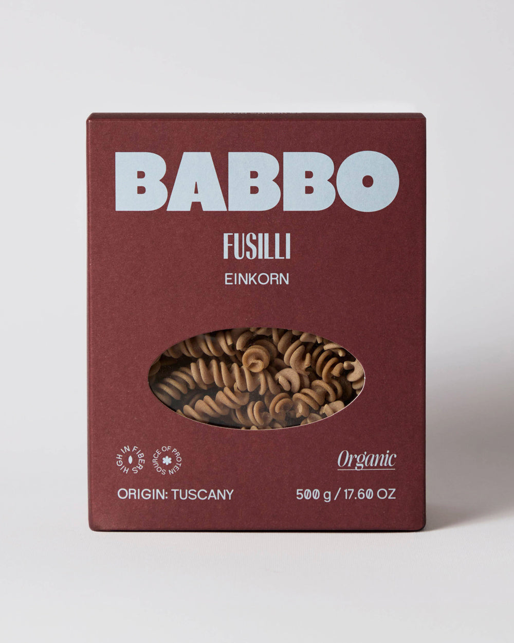 A 500g box of Babbo Fusilli Einkorn pasta, featuring a dark reddish-brown box with white text and a clear oval window showing the brown pasta inside, set against a neutral light gray background. Available via the Pantry Collection from Wildschut Antiques.