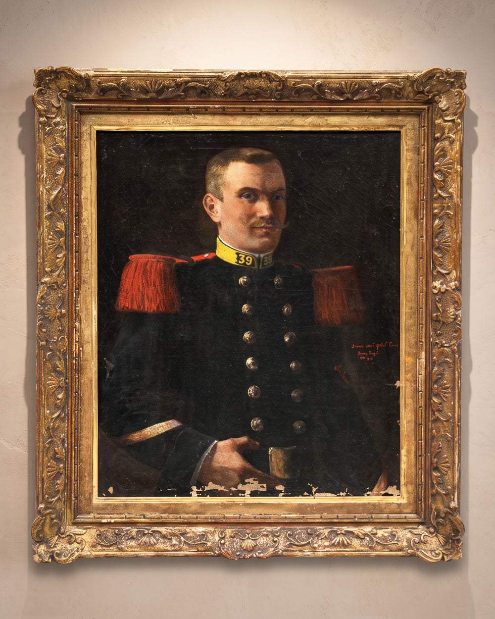 A framed portrait oil painting of a man in a dark military uniform with red shoulder epaulets and gold buttons, set in an ornate carved gold frame and hanging on a light beige wall. Available via Wildschut Antiques.