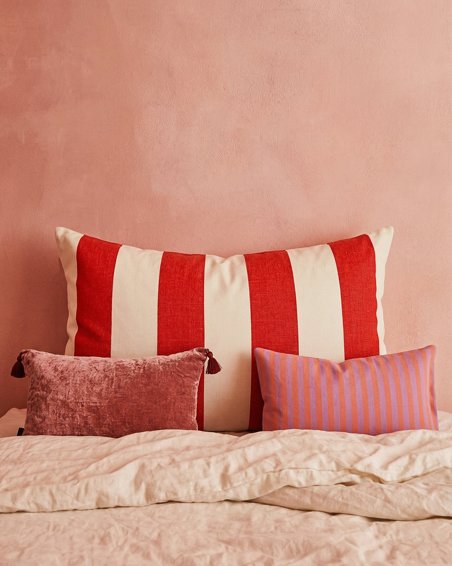 Three decorative pillows with red and white stripes, pink texture, and pink and purple pattern on a bed against a pink wall.