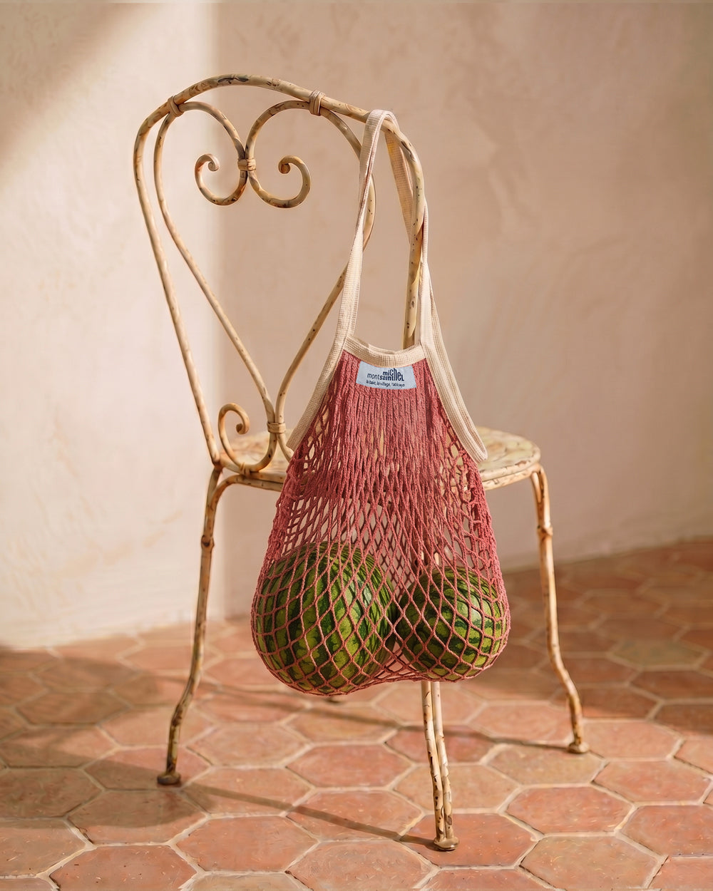 Pink mesh bag with green watermelons on a decorative vintage French metal chair against a beige wall. By Filt 1860, available at Wildschut Antiques.