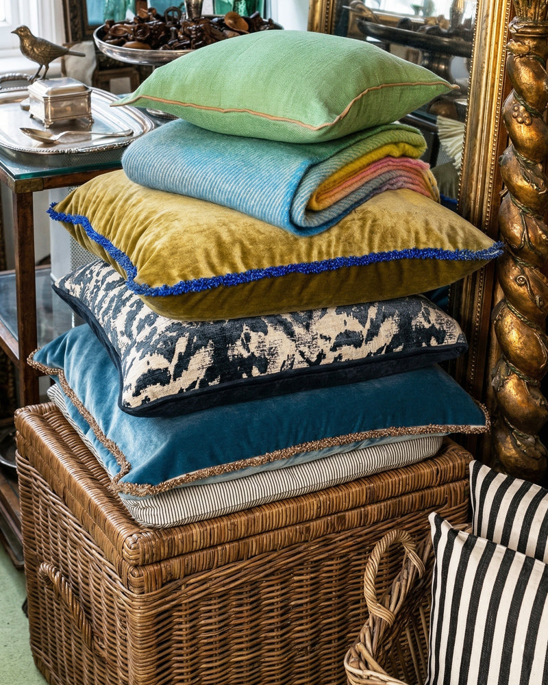 Stack of colorful pillows on a wicker basket with decorative items in the background, in store at Wildschut Antiques.