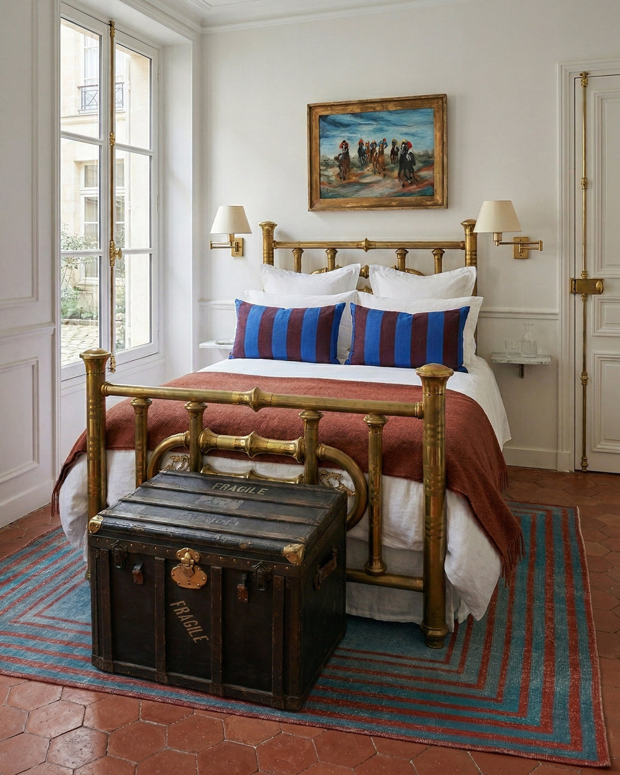 Classic bedroom with a brass bed, wooden trunk, and blue and red striped rug, all items available via Wildschut Antiques.