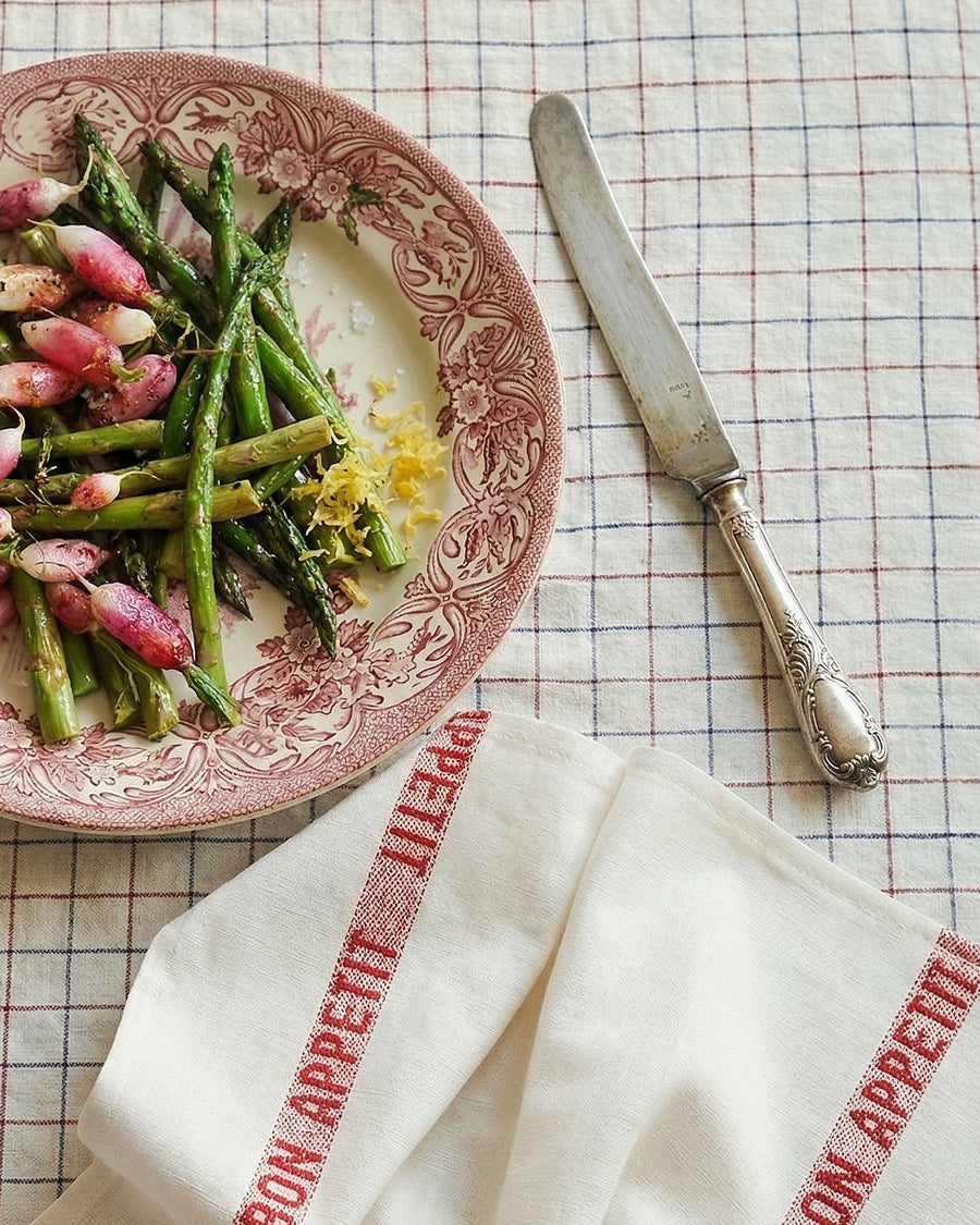 Plate of asparagus salad with radishes on a checkered tablecloth, accompanied by a knife and a linen napkin with 'Bon Appetit' text in red, by Charvet Editions available via Wildschut Antiques..