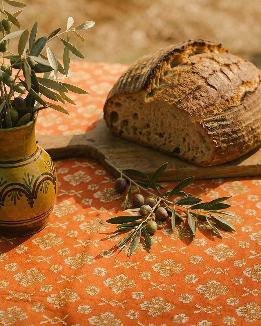 Loaf of bread on a wooden board with an olive branch and pot on an orange floral block printed tablecloth, available via Wildschut Antiques.