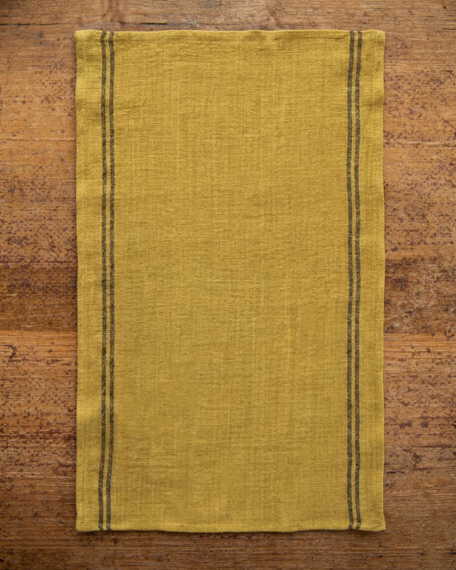 Ochre yellow linen towel with dark stripes on a wooden surface. Linen country towel by Charvet Editions available via Wildschut Antiques.