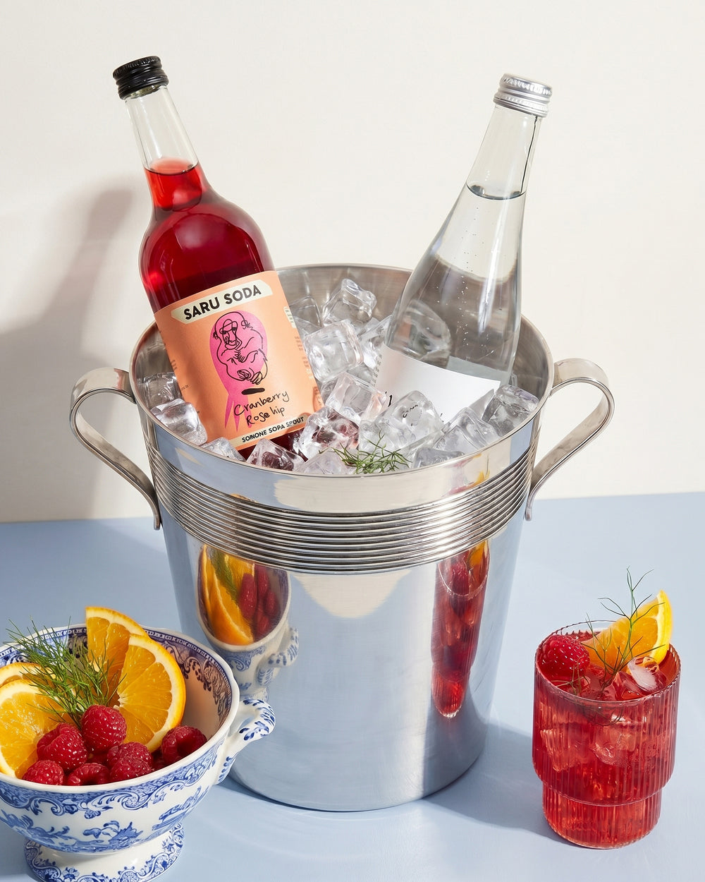 Bottle of Saru Soda Lemonade and a bottle of sparkling water in a vintage silver plated ice bucket with a glass of lemonade and a bowl of fruits on a light blue background. Available at Wildschut Antiques.