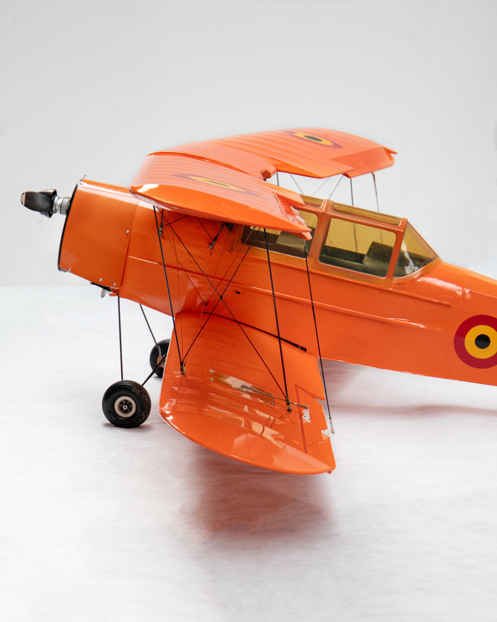 An orange model high-wing biplane with a propeller, shown from a three-quarters rear angle on a light gray studio background.