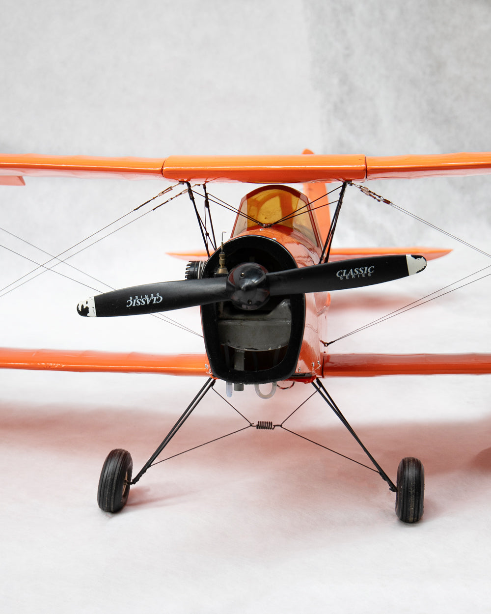 An orange and black model biplane with a black propeller, shown from a low front angle against a light gray background.