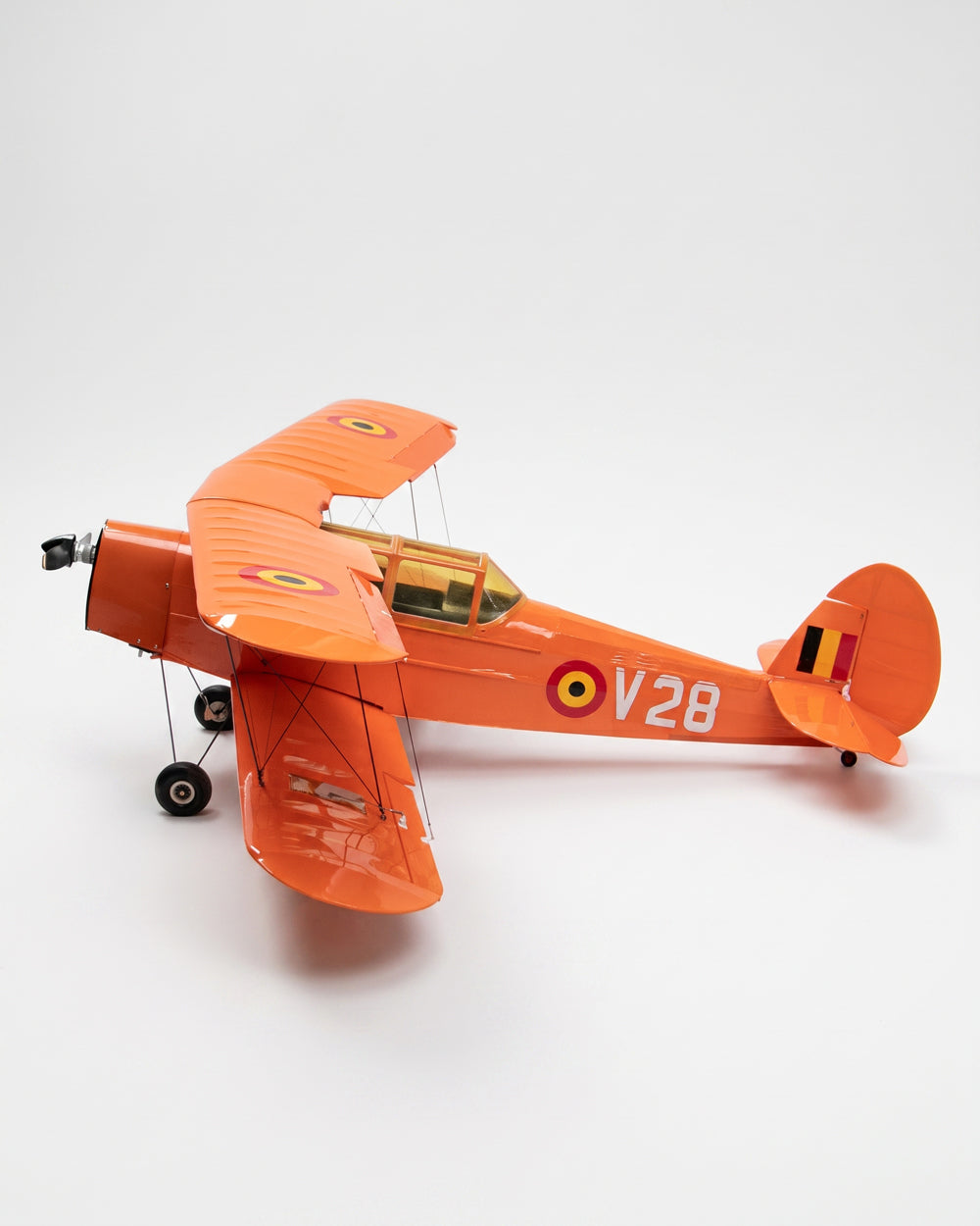 An orange model biplane with the markings 'V28' and a small Belgian flag on the tail, photographed on a seamless light gray studio background.