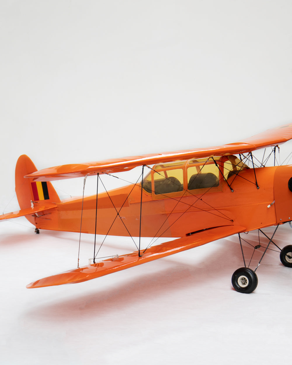 An orange model biplane with a transparent canopy, black wire struts, and black wheels, photographed on a seamless white studio background.