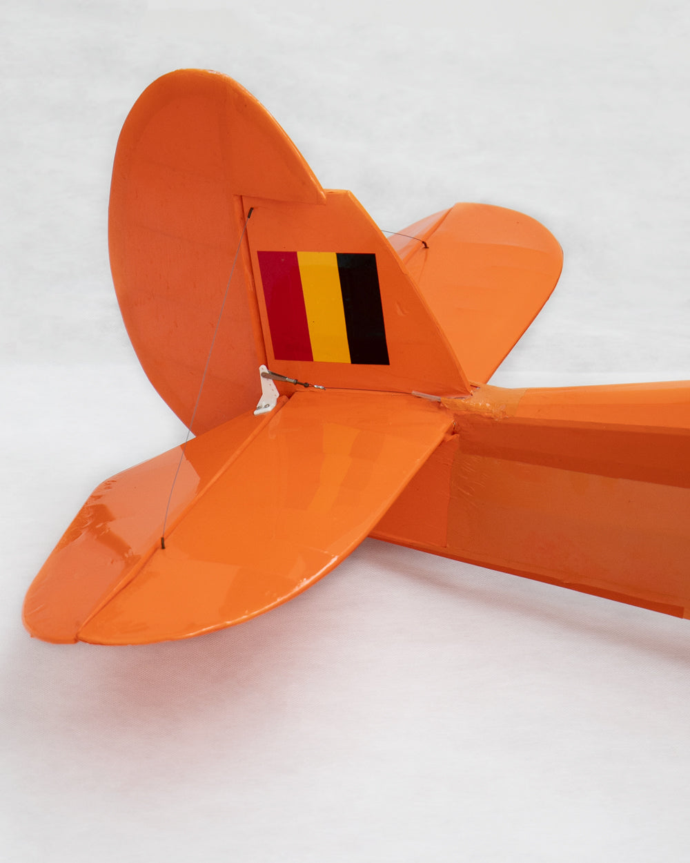 An orange model airplane tail section with a small vertical flag detail in red, yellow, and black, photographed on a light gray background.