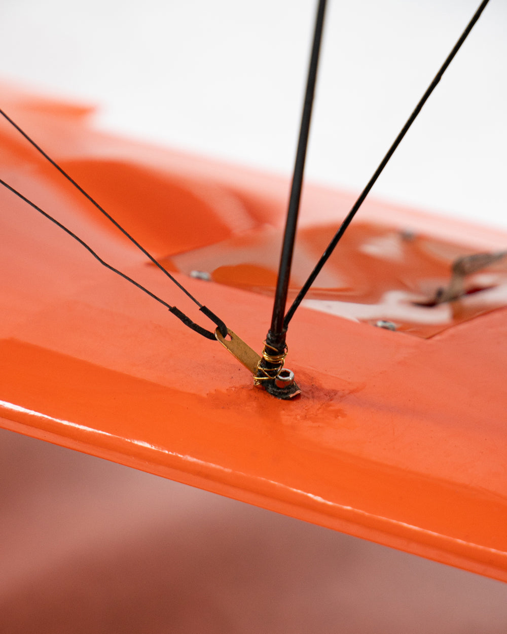 Close-up of an orange airplane model wing with black suspension cables threaded through a metal fitting and secured with gold wires.
