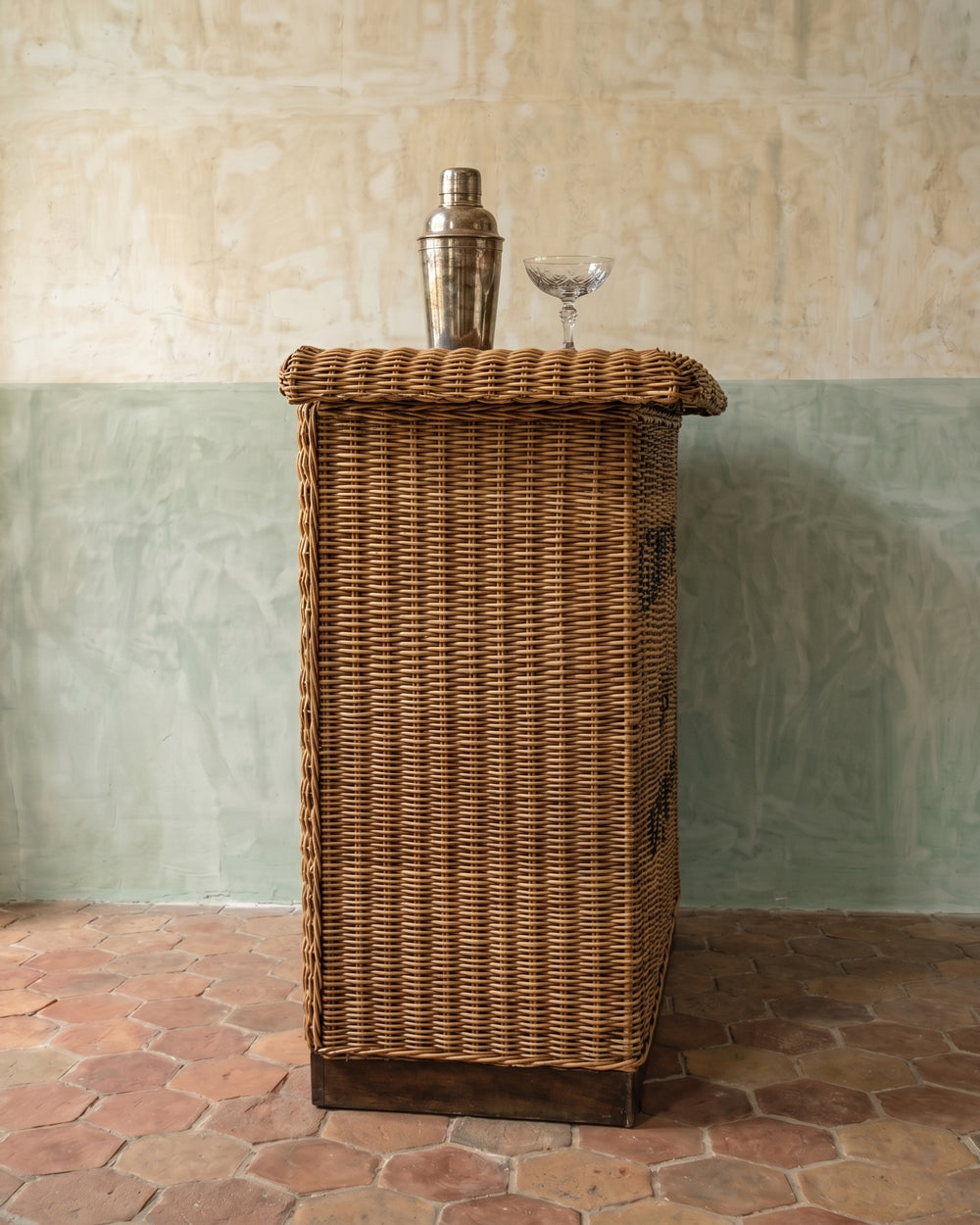 Wicker bar cart against a textured wall with a cocktail shaker and glass on top.