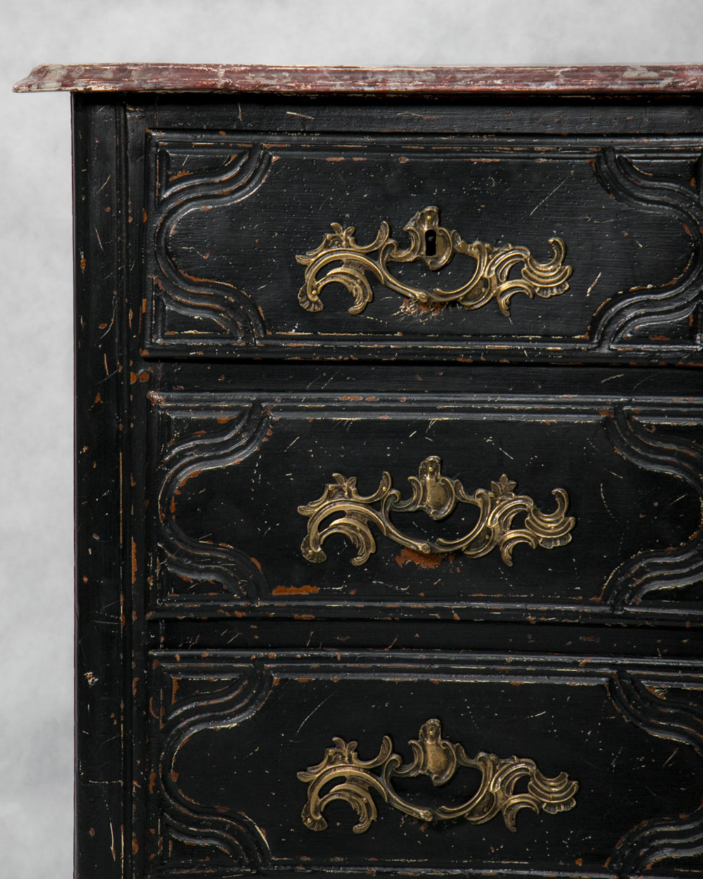 Close-up of an antique black wooden chest of drawers with ornate gold-tone metal drawer pulls and a worn reddish-brown top surface, photographed against a light gray background.