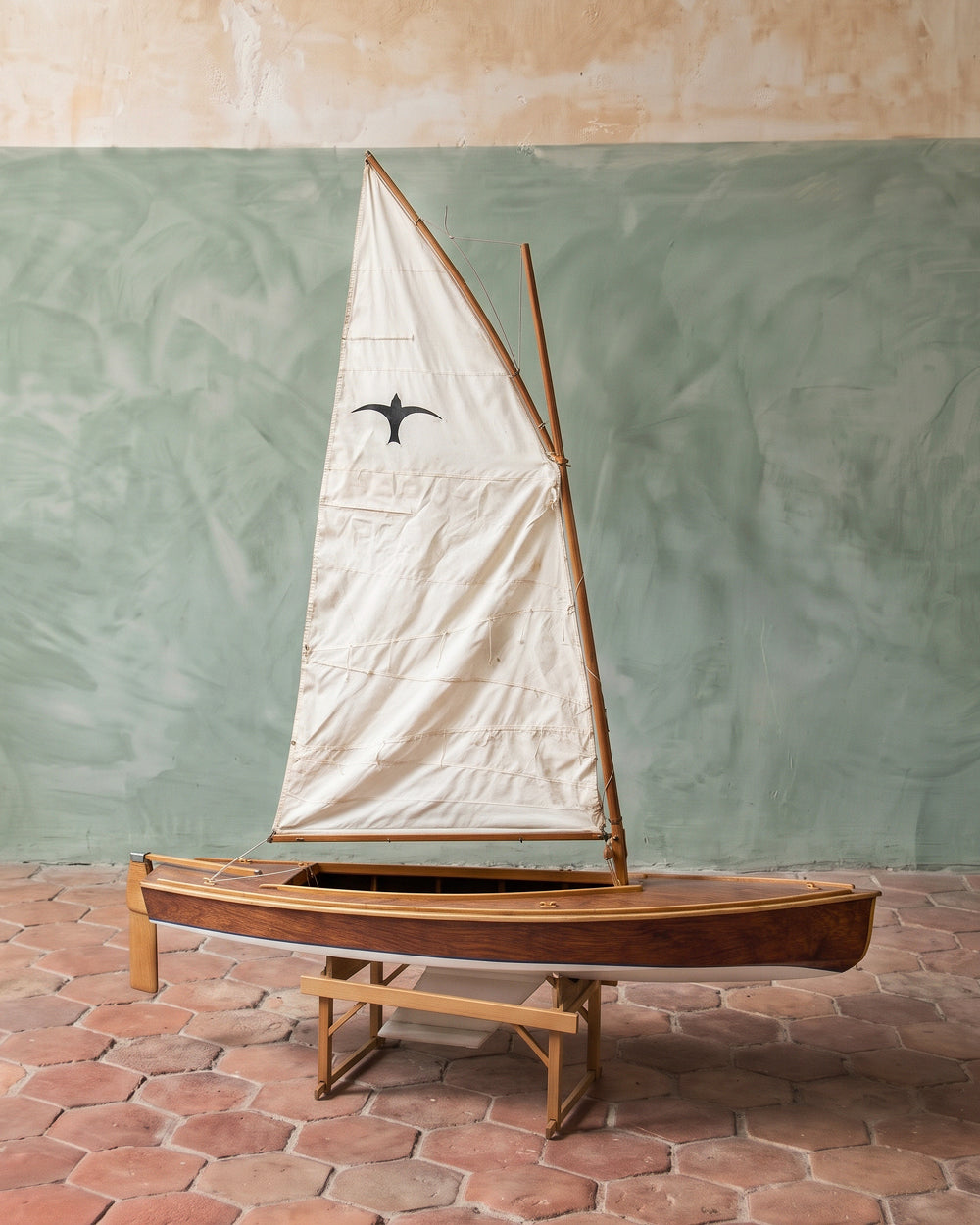 A vintage wooden Valk model sailboat with a cream-colored canvas sail featuring a black bird emblem, displayed on a wooden stand against a textured green and beige wall. Available via Wildschut Antiques.