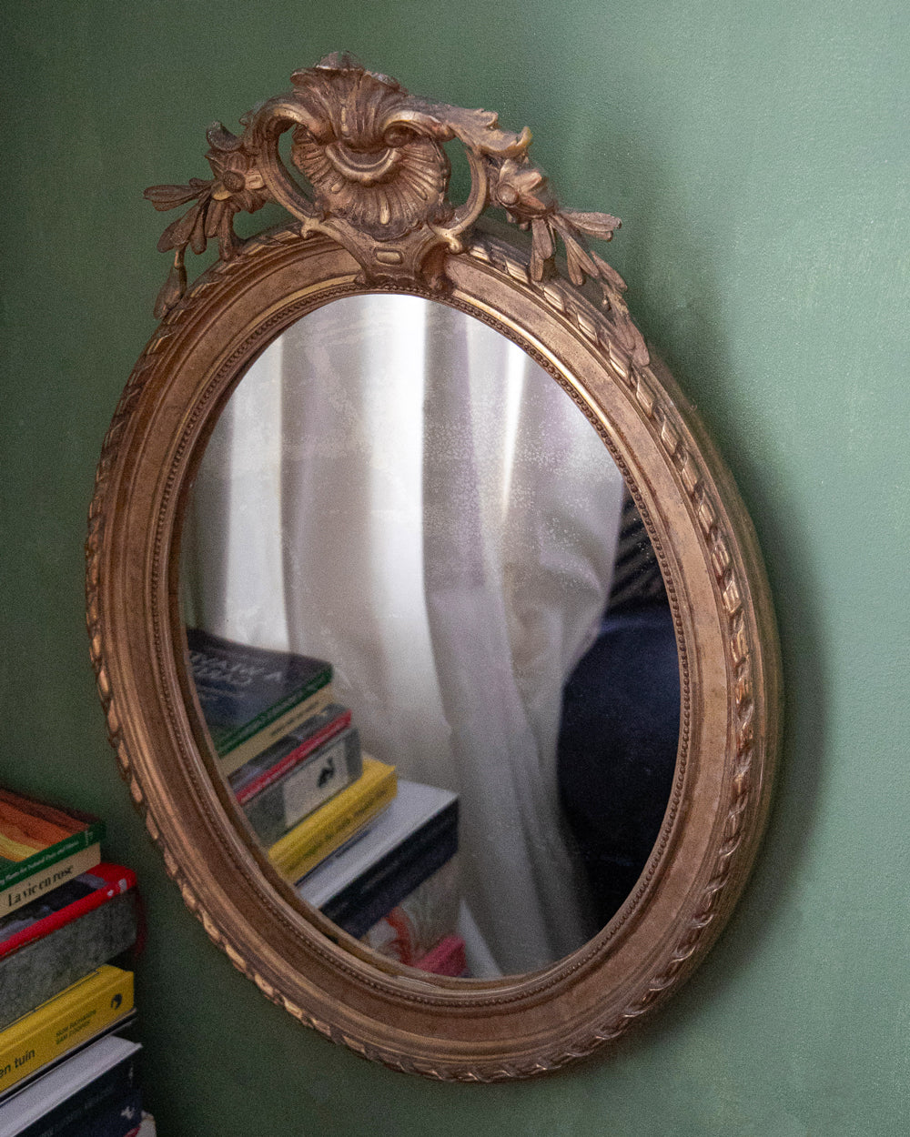 An ornate oval wall mirror with a carved gold-toned frame, mounted on a textured green wall. The reflection shows white curtains and a stack of books, and a matching stack of books is visible in the bottom-left corner of the frame.